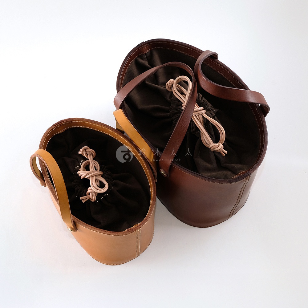 兩只職人手作皮革束口手提包，一深棕色，一淺棕色。皆為橢圓形狀，配有束口內袋及皮革提把。