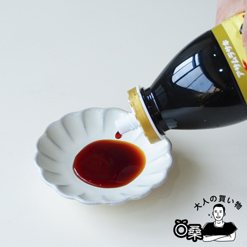 一隻手正握著朝紫品牌廣島牡蠣醬油瓶，將深褐色的醬油緩慢倒入白色花瓣造型的陶製小碟中，畫面右下角帶有大人的買い物與桑的標誌圖樣，整體呈現簡約的生活感視角。