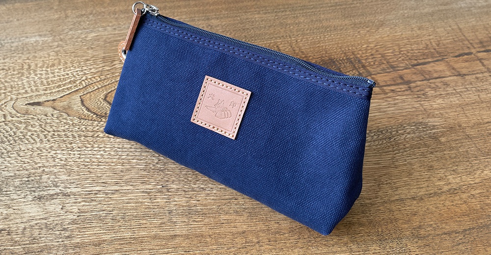 深藍色石洗帆布製成的小型收納包，附有拉鍊及一個皮質品牌標籤，放置在木紋桌面上。