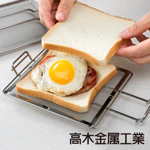銀色不鏽鋼網烤吐司夾，夾著兩片吐司、培根和煎蛋，另一片吐司由雙手持握準備蓋上。