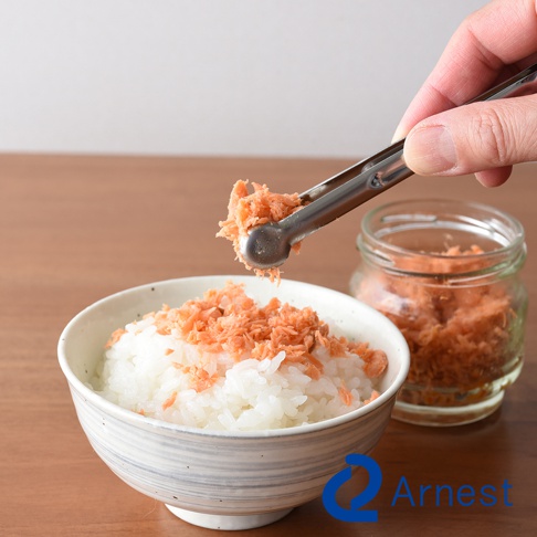 逸品物創不鏽鋼萬用料理夾，夾起鮭魚鬆，準備放到裝有白米飯的碗裡，背景是裝有鮭魚鬆的玻璃罐。