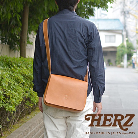 日本HERZ職人手作皮革軟皮橫式斜背包，由側面看，一名男子穿著深藍色襯衫，搭配米白色長褲，斜背著焦糖色皮革斜背包。