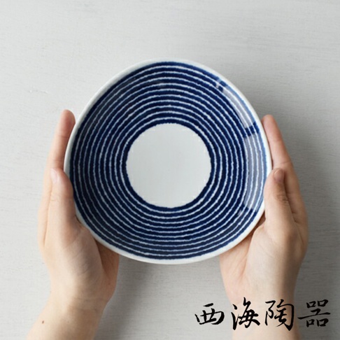 一雙手捧著一個白色陶瓷盤，盤面繪有深藍色同心圓紋路，紋路向外擴散，盤中央為純白色圓形區域。