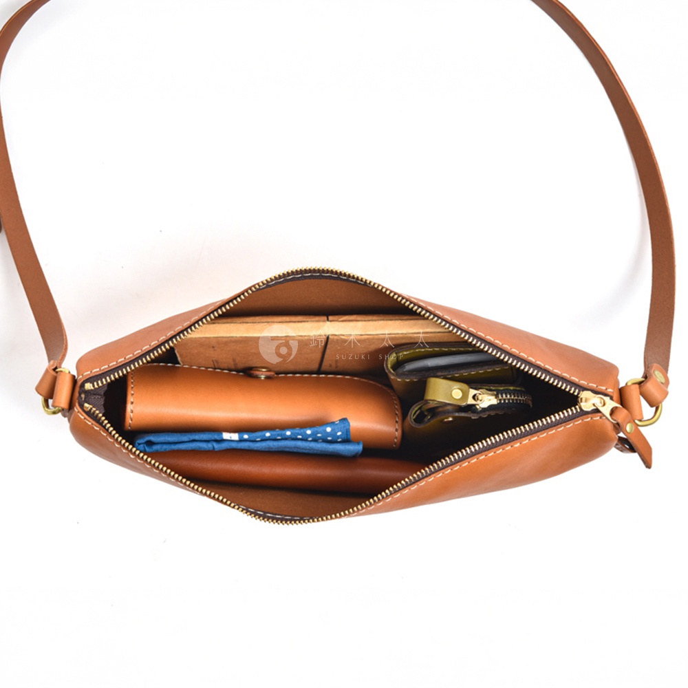 日本HERZ職人手作皮革半圓馬鞍包，棕色皮革材質，內部有長形皮夾、藍色方格布、筆狀物及零錢包等物品，銀色拉鍊，一條細長的皮革揹帶。