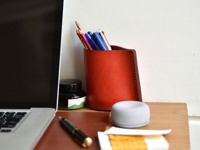 橘紅色皮革製成的手工筆筒，內有彩色鉛筆、原子筆和鋼筆，旁邊擺放著墨水瓶、筆記本和灰色小物。