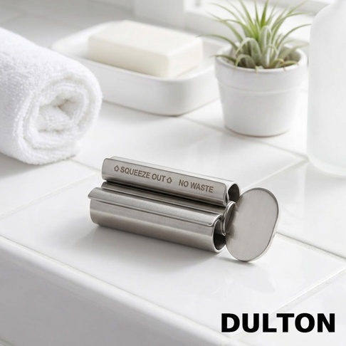 放在白色磁磚平面上的DULTON不鏽鋼牙膏擠壓器，金屬材質呈現霧面光澤，機身刻有SQUEEZE OUT NO WASTE字樣，旁邊放置著捲起的白毛巾、肥皂盒與一盆小型多肉植物。