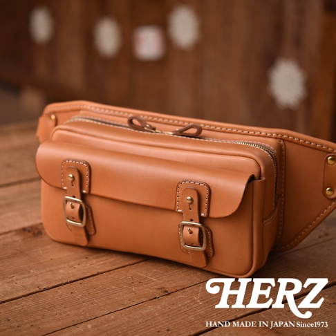 職人手作皮革騎士隨身斜背包，採用光滑的淺棕色皮革製成，配有銅色釦環和拉鍊，包款設計簡潔，適合隨身攜帶。