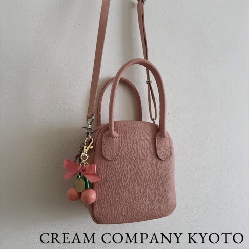 粉紅色荔枝紋皮革手提包，配有可拆卸肩帶及櫻桃造型吊飾，整體風格簡約可愛。