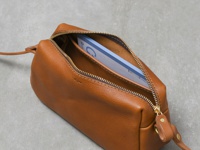 淺棕色皮革製的職人手作輕巧拉鍊單肩包，拉鍊部分採金色金屬材質，包內可見一本淺藍色筆記本。