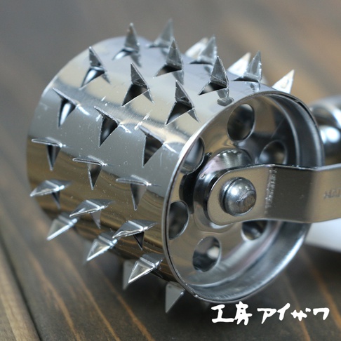 不鏽鋼滾輪斷筋器，圓筒狀，表面佈滿三角形尖刺，滾輪兩側有圓形孔洞，整體為金屬銀色，置於木紋背景上。