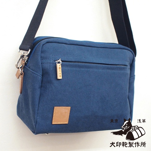 藍色帆布斜背包，正面有一個拉鍊口袋，附有木質吊飾，下方有一個皮質標籤，右下角印有日文圖案與文字。