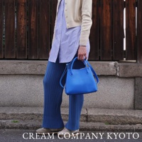 一位身穿藍色條紋襯衫、米色開襟毛衣和藍色寬褲的女士，手提一個亮藍色皮革托特包，包包有一條可拆卸肩帶。