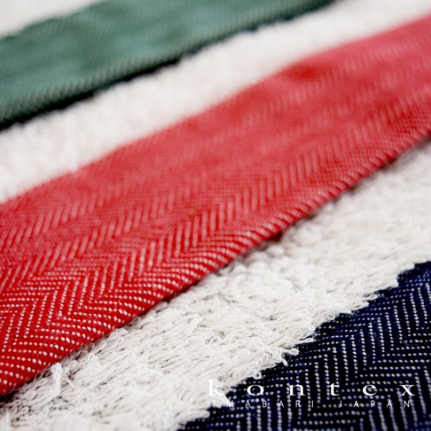 HERRINGBONE 今治長纖有機棉斜紋浴巾，採用多色條紋設計，包含紅色、白色、綠色和海軍藍。浴巾表面可見經典的斜紋編織紋理，以及柔軟的絨毛質地，展現其高品質的棉質材質。