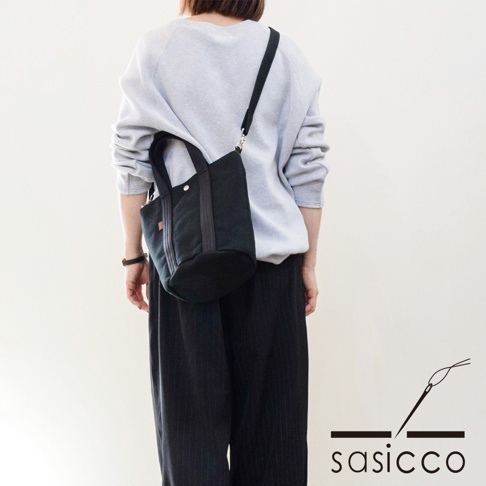 一位女性身著淺灰色衛衣和黑色條紋長褲，側背一個黑色三河木綿刺子織兩用水桶包。包包為圓筒狀，有兩個短提把和一條斜背帶。