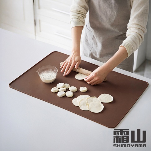 日本霜山長方形矽膠不沾黏揉麵墊，一個人正在廚房使用擀麵棍擀壓麵皮，旁邊放著一碗麵粉和揉好的小麵團。