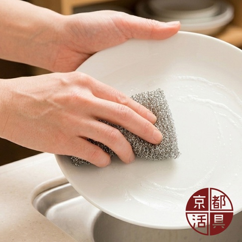 一雙手正拿著京都活具的不沾鍋專用菜瓜布，在水槽上方清潔白色餐盤，菜瓜布呈現獨特的金屬色澤與網狀編織質感，特寫角度展示了其在處理廚房髒污時的實際操作情境，右下角印有京都活具的品牌標誌。
