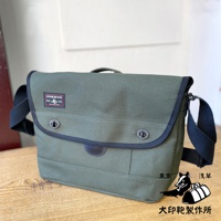 這款黑犬印純棉帆布雙扣郵差包，採用軍綠色帆布材質，配有深色滾邊與雙扣設計。包包有可調整的肩帶，正面有一個紅色標籤，上面印有日文文字與狗狗圖案。