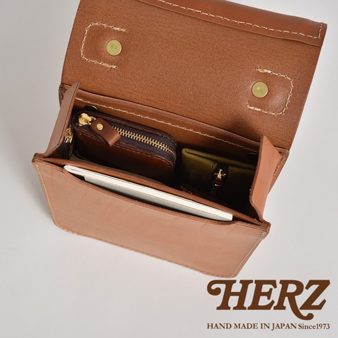 職人手作淺棕色皮革立體口袋兩用單肩包，內部有深棕色拉鍊零錢包和軍綠色小物包，以及一本白色筆記本。