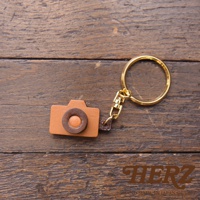 職人手作皮革相機造型鑰匙圈，主體為淺棕色皮革，鏡頭部分為深棕色皮革點綴，搭配金色金屬鑰匙圈，整體精緻可愛。