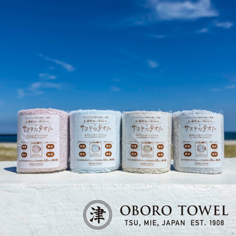 四條捲起的毛巾，分別為淡粉色、淺藍色、米白色和淺灰色，皆展示產品包裝上的日文說明，背景為藍天和海灘。