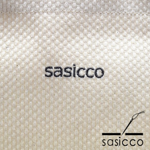 三河木綿刺子織經典托特包的材質特寫，展示米白色織物表面精緻的方形紋理，以及黑色刺繡的品牌標誌「sasicco」和針線圖示。