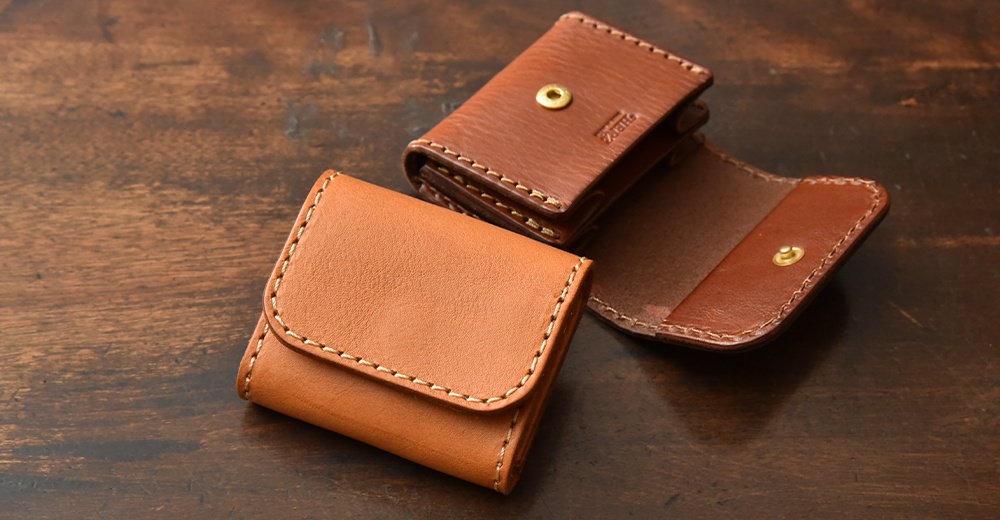 桌面上放置兩個日本HERZ品牌的手作皮革零錢包，左側為關閉狀態呈現淺棕色，右側為打開狀態呈現深棕色，展示其皮革紋理、縫線細節以及金屬鈕扣設計。