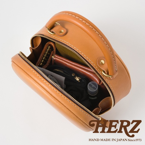 HERZ 職人手作皮革兩用圓形包，展示包內收納，有卡夾、皮夾、手機和瓶裝水，品牌 LOGO 與日製標語印製在右下角。