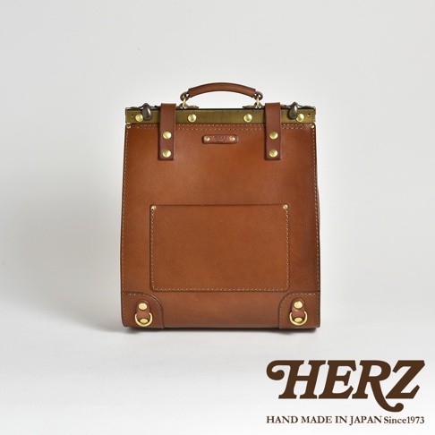 職人手作皮革復古口金後背包，以棕色皮革製作，設有金屬扣環與鉚釘裝飾，正面有一個方形貼袋。