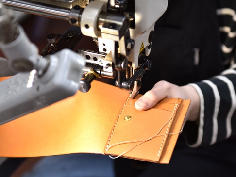 職人以縫紉機正在製作一個淺棕色皮革的直式卡片收納雙折短夾，皮革上有精緻的縫線與金屬釦。
