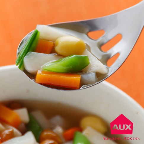 銀色不鏽鋼濾水菜匙，盛著切塊的胡蘿蔔、白蘿蔔、毛豆和黃豆，湯汁從匙孔中瀝出，背景為橙色。