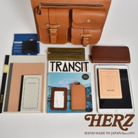 職人手作皮革三口袋三用後背包，展示多種搭配小物，包括筆記本、雜誌、證件套、鑰匙包、錢包及平板電腦。後背包為皮革材質，顏色為天然棕色，配有兩個前袋，袋蓋皆有金屬扣件及拉鍊，整體呈現簡約質感。