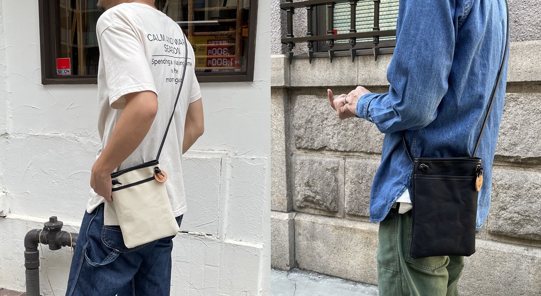 兩名男子展示 uncle 高島帆布皮革散步包，其中一人揹著米白色帆布包，另一人揹著黑色帆布包，兩款包皆搭配皮革飾邊與細肩帶，並有圓形皮革吊牌。