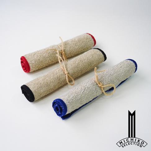 自在道具品牌標誌旁，有三條捲成長條狀的棉麻質地擦拭巾，分別以紅色、黑色和藍色收尾，並各自以麻繩綁住，置於白色背景上。
