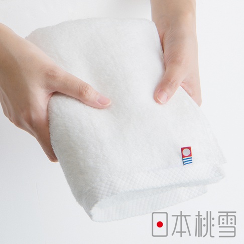 雙手捧著一條純白色今治超長棉浴巾，浴巾質地蓬鬆柔軟，邊緣有精緻的菱格紋縫線，角落處有一個藍白紅三色的小標誌，下方印有日文「日本桃雪」。