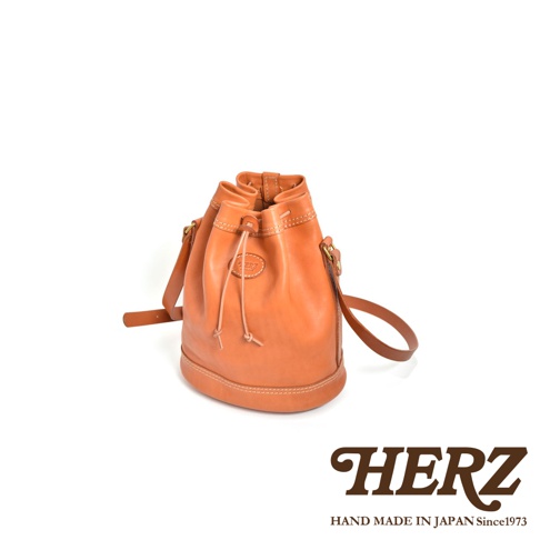 職人手作皮革束口單肩包，橘棕色皮革，束口設計，配有可調式肩帶，包身圓潤，以白色背景呈現。