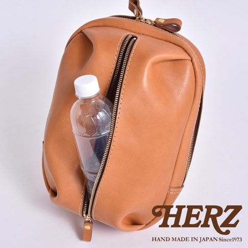 日本HERZ手工皮革立體運動斜背包，棕色牛皮革材質，拉鍊開口設計，側邊可見放入寶特瓶，左下方有HERZ品牌標誌與HAND MADE IN JAPAN Since1973字樣。