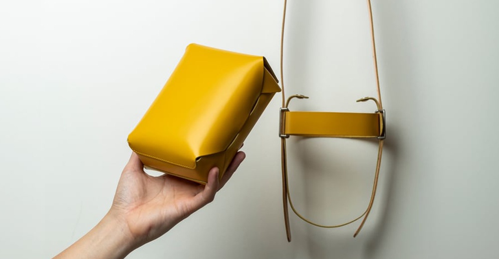 FABRIK 皮革可拆式多用途迷你斜背包，展示一個亮黃色的迷你斜背包，由手持，旁邊懸掛著帶有金屬扣環的包帶