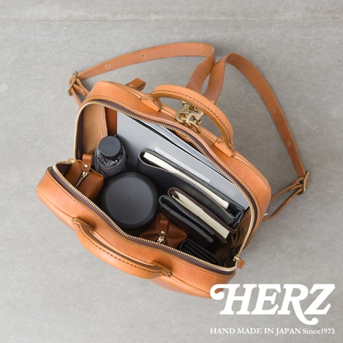 職人手作皮革Organ商務後背包，打開內部陳列著筆記本、文件夾、折疊傘、圓形收納盒及皮夾等物品，展現實用收納空間。