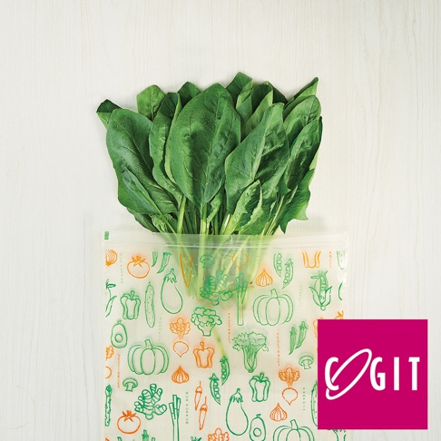 一疊新鮮的綠葉蔬菜（菠菜）置於一個半透明的環保鮮綠袋中，袋身印有綠色和橘色的蔬果圖案，如番茄、辣椒、南瓜、玉米、花椰菜等。