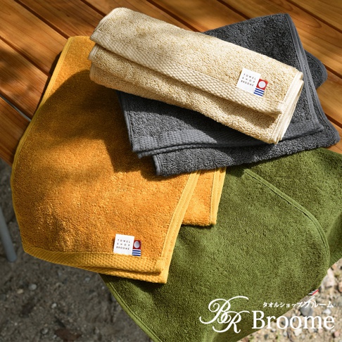 黃色、灰色、卡其色和綠色的今治純棉毛巾，部分疊放於木桌上，毛巾上有品牌標籤。