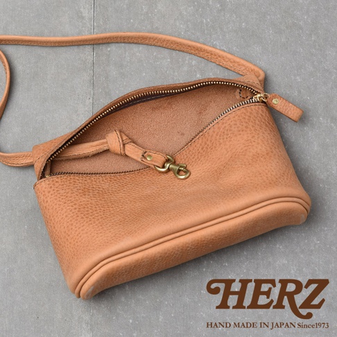 HERZ 品牌標誌，下方為 HAND MADE IN JAPAN Since1973 字樣。 一個淺棕色的皮革斜背包，背包的拉鍊敞開，露出內裡。背包帶和金屬扣環清晰可見。
