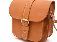 棕色皮革單肩包，包蓋上有圓形十字繡裝飾，配有黃銅搭扣。包身側面縫線精緻，整體呈現簡約復古風格。