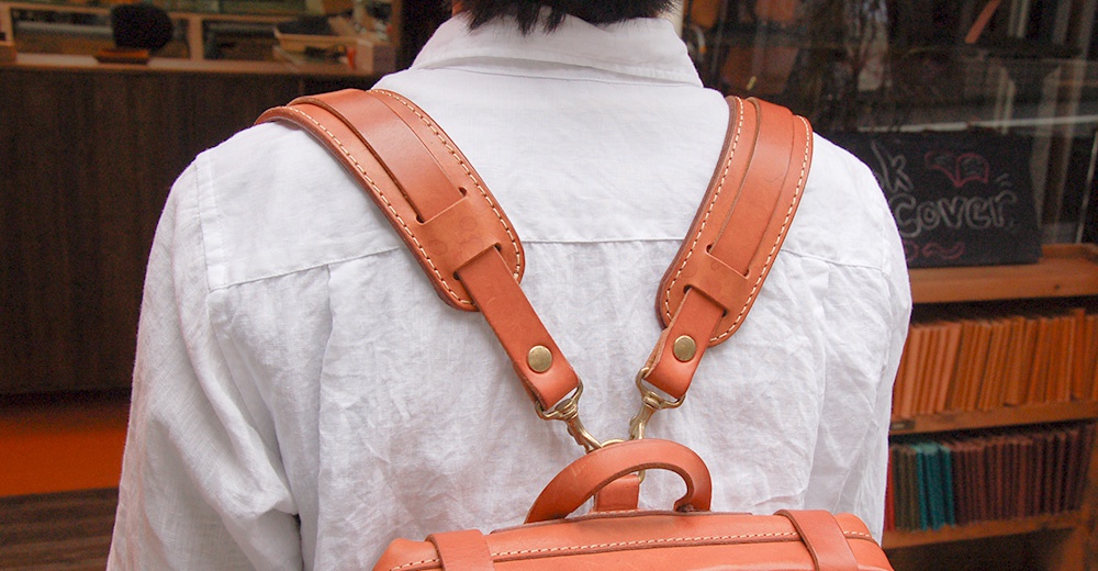 職人手作皮革Big肩墊，呈現自然棕色，皮革材質，雙肩背帶設計，配有金屬扣環，與白色襯衫搭配。