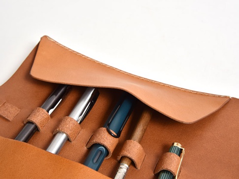 淺棕色皮革捲捲收納筆袋，內部有五個皮革筆環，可固定黑色、銀色、藍綠色、木質及綠色條紋的筆。