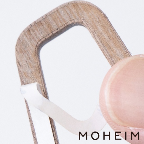 展示一個木製標示牌的局部特寫，邊緣有細緻的木紋，牌面為淺棕色。一隻手指正將背面的白色黏膠剝離，準備安裝。