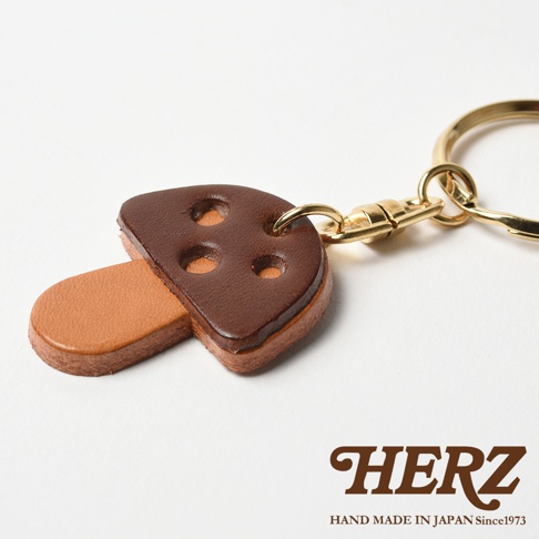 職人手作皮革香菇造型鑰匙圈，傘蓋為深棕色皮革，有三個圓孔裝飾；菌柄為淺棕色皮革，造型簡約可愛，搭配金色金屬掛環與扣環。
