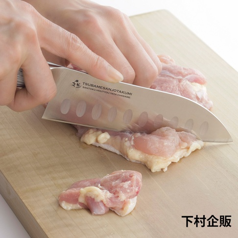 鉬釩不鏽鋼三德刀，刀刃上有橢圓形凹槽，正在切塊的粉色生雞肉，置於淺木色砧板上。