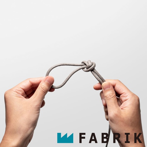 FABRIK 品牌標誌旁，雙手正將一條灰色繩子打成一個活結，繩子的另一端垂下。