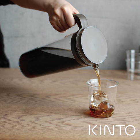 手持深棕色把手的透明玻璃冷泡壺，正將深色液體倒入裝有冰塊的玻璃杯中。