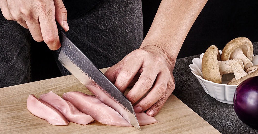 一名廚師用一把日式手工刀將生雞胸肉切片，旁邊還有香菇和茄子